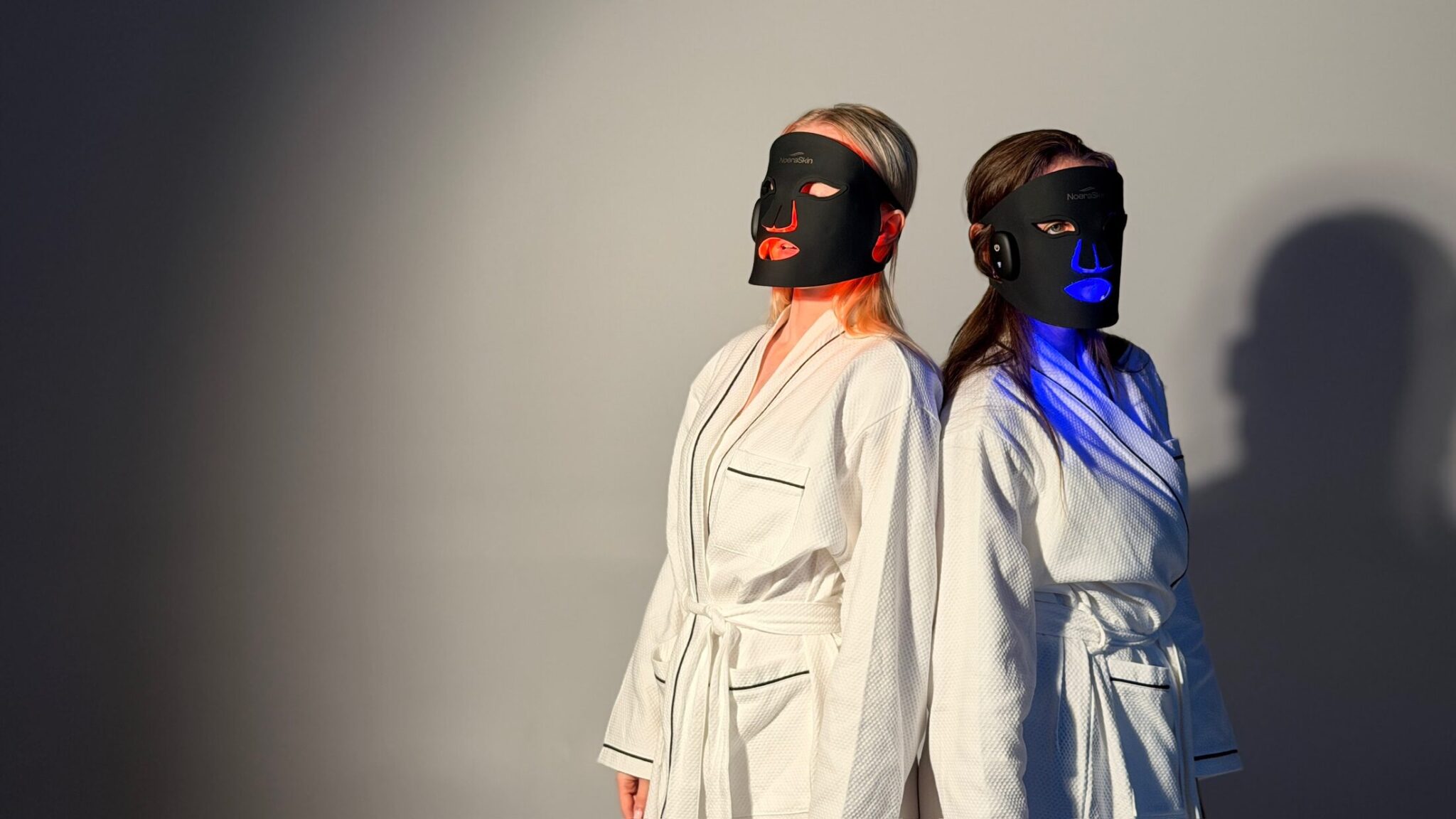 Two women in white robes, back to back, wearing the mask