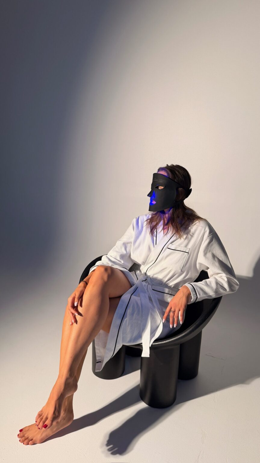 A woman in a white robe seated in a black chair wearing the mask